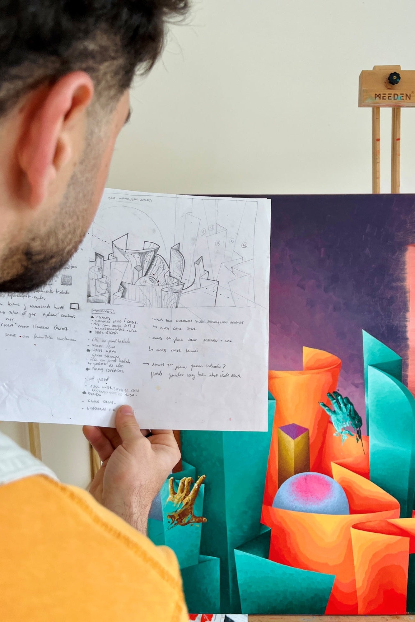 Person holding a drawing next to a colorful art piece on an easel